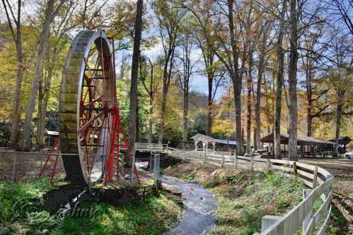 A large water wheel beside a stream, surrounded by trees with autumn foliage and a rustic shelter in the background.