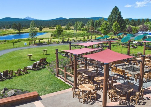 A scenic outdoor area with wooden tables, chairs, and umbrellas, overlooking a pond and mountains in the background.