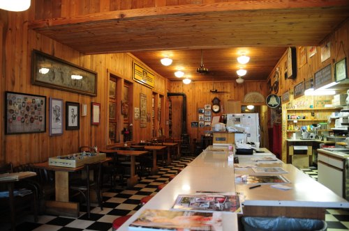 Cozy diner interior with wooden walls, checkered floor, tables, and vintage decor. Brightly lit with a welcoming atmosphere.