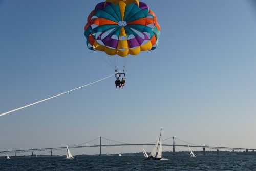 Two people parasailing above a body of water, with a colorful parachute and a bridge in the background.