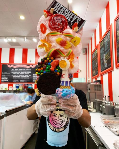 A towering dessert with ice cream, candy, cookies, and whipped cream, held by a person in a shop with red-striped walls.