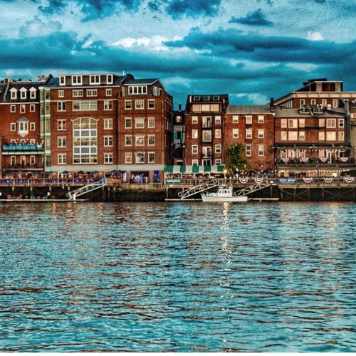 A waterfront view of brick buildings with balconies, reflecting in the water under a cloudy sky.