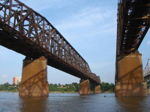 A view of a large, rusty metal bridge spanning over a river, with concrete pillars and a clear blue sky above.