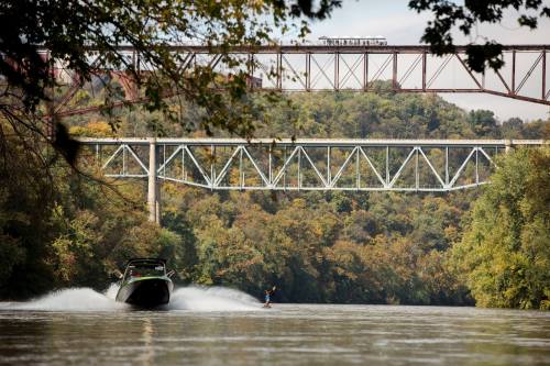 A boat pulls a water skier on a river, with two bridges and lush trees in the background.