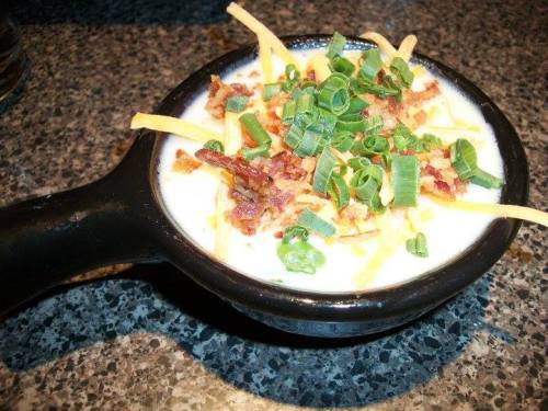 A black bowl filled with creamy soup topped with cheese, bacon bits, and chopped green onions on a granite surface.