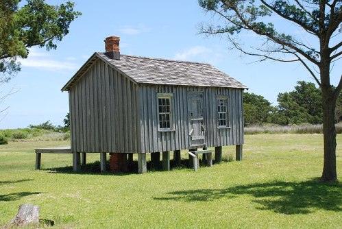 A small, weathered wooden house on stilts, surrounded by grass and trees under a clear blue sky.