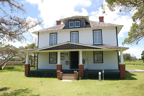 A two-story white house with a porch, surrounded by grass and trees under a blue sky with clouds.
