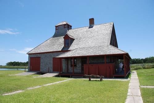 A large, historic house with a gray shingle roof and red accents, surrounded by green grass and a clear blue sky.