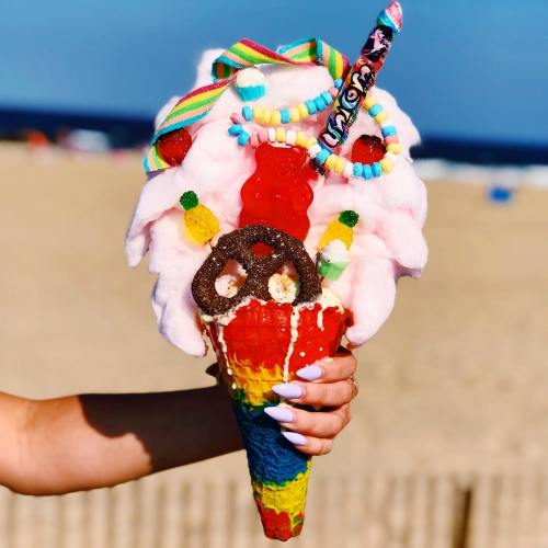 A colorful ice cream cone topped with cotton candy, candy pieces, and a rainbow cone, held against a beach backdrop.