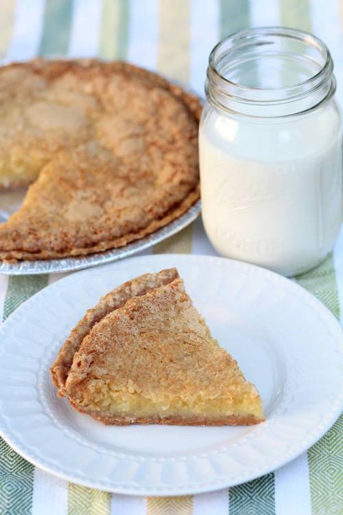 A slice of pie on a plate next to a jar of milk, with a whole pie in the background on a striped tablecloth.