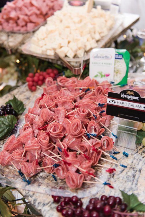 A platter of rolled salami skewers, cheese cubes, and grapes on a marble table, decorated with greenery.