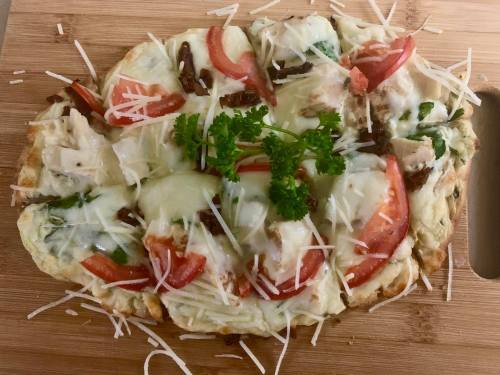 A flatbread topped with cheese, tomatoes, greens, and herbs, served on a wooden cutting board.