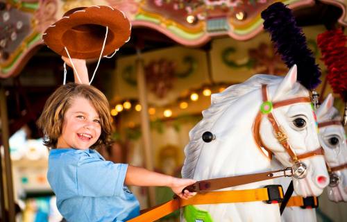 A smiling child in a blue shirt rides a white carousel horse, holding a cowboy hat aloft.