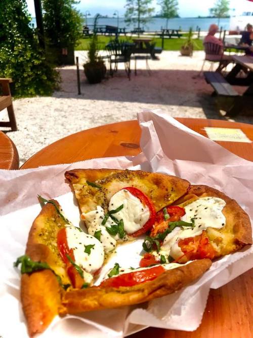 A slice of pizza topped with tomatoes, mozzarella, and herbs, set on a wooden table with a scenic outdoor view.