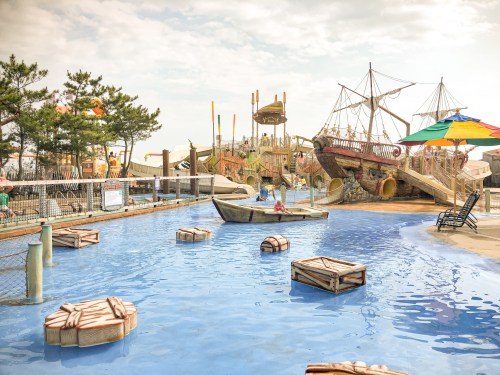 A water park scene featuring a pirate ship, a pool with floating crates, and colorful umbrellas under a cloudy sky.