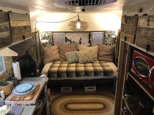 Cozy interior of a van with a daybed, decorative pillows, wooden accents, and a small kitchen area.