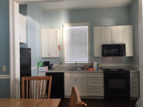 Bright kitchen with white cabinets, black appliances, and a window letting in natural light. Wooden dining table in foreground.