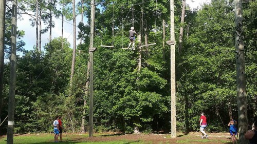 A person climbs a high ropes course while others observe from the ground in a wooded area.