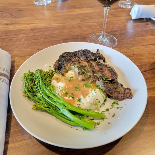 A plate of grilled steak with mashed potatoes, topped with gravy, and a side of sautéed greens, accompanied by a glass of red wine.