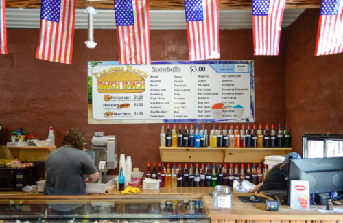 A snack shack interior with a menu board, colorful syrup bottles, and American flags. Two people are working behind the counter.