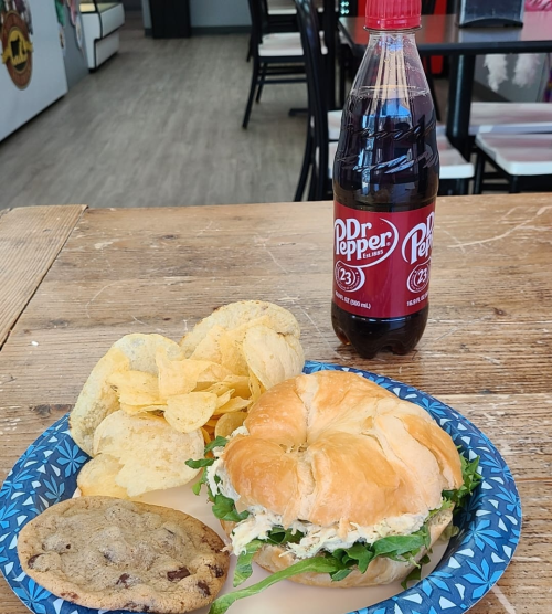 A plate with a croissant sandwich, chips, a cookie, and a bottle of Dr Pepper on a wooden table.
