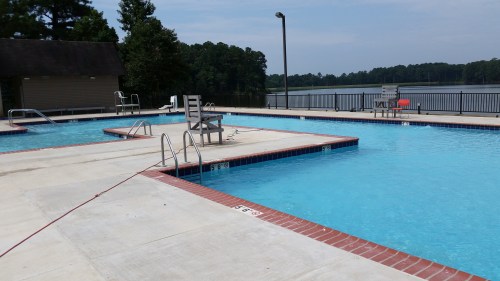 A serene outdoor pool area with multiple sections, surrounded by trees and a lake in the background.