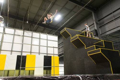 A person jumps off a high platform at a trampoline park, while another person stands on the platform above.