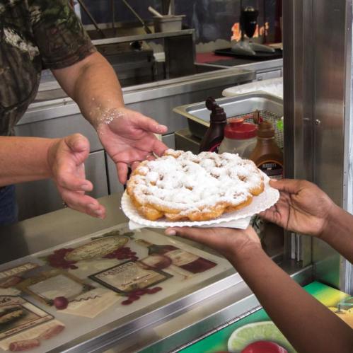 A hand reaches out to receive a plate of funnel cake dusted with powdered sugar at a food stand.