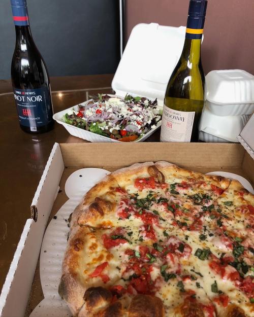 A pizza with tomatoes and herbs, two bottles of wine, and a salad in takeout containers on a table.