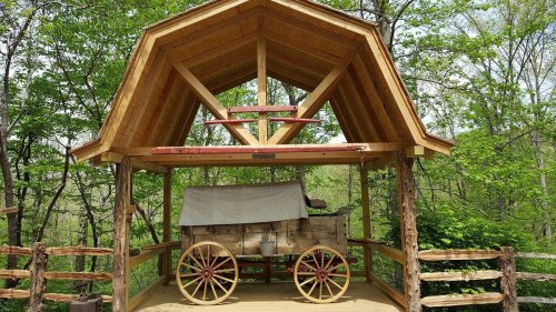 A wooden shelter with a wagon underneath, surrounded by lush green trees.