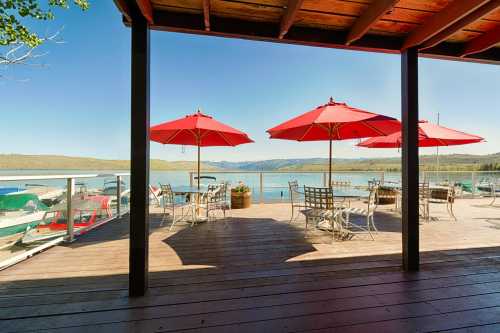 A lakeside deck with red umbrellas over tables, overlooking boats and calm water under a clear blue sky.