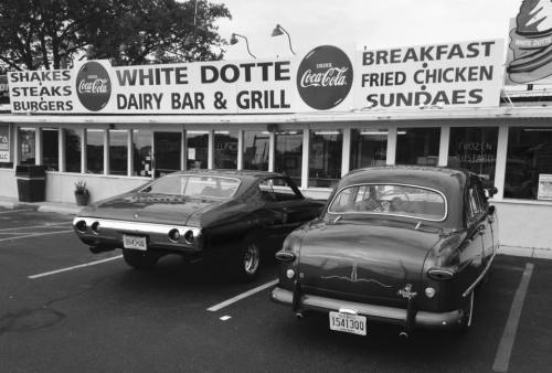 Two classic cars parked in front of a retro diner, "White Dotte Dairy Bar & Grill," featuring a vintage sign.