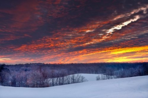 A winter landscape with snow-covered hills and a vibrant sunset illuminating the cloudy sky.