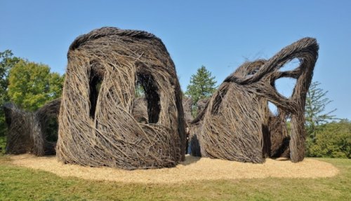 A large, abstract structure made of intertwined branches, set in a grassy area with trees in the background.