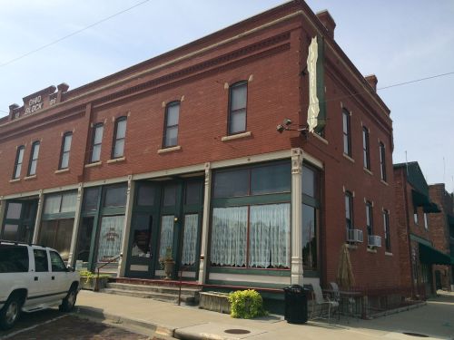 A red brick building with large windows and white curtains, featuring a green awning and outdoor seating area.