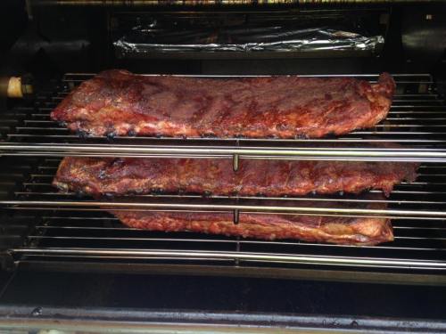Two racks of ribs cooking on a grill, with a smoky glaze and a rich brown color, placed on metal racks.