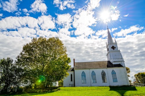 A white church with a tall steeple stands beside a large tree under a bright sun and cloudy blue sky.