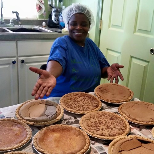 A smiling woman in a hairnet presents a table of freshly baked pies in a kitchen setting.
