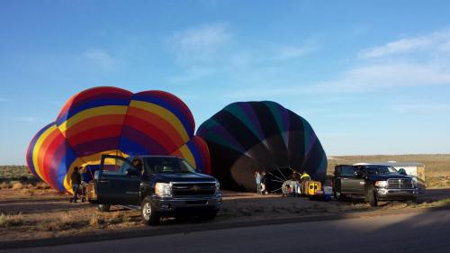 Two colorful hot air balloons being prepared for launch next to two pickup trucks in a desert landscape.