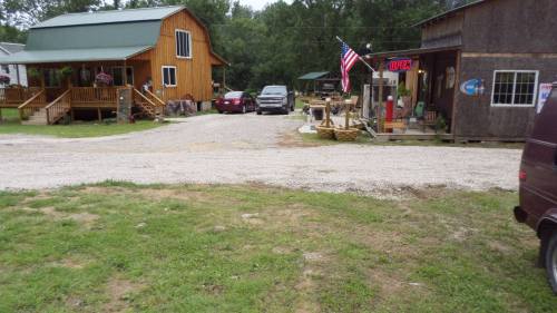 A gravel road leads to wooden buildings, one with an "OPEN" sign and an American flag, surrounded by greenery.