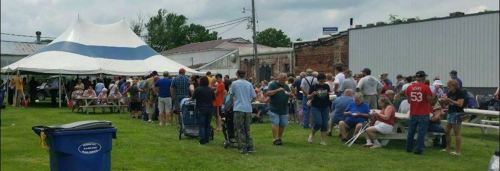 A large crowd gathers outside under a tent, with people in line and enjoying food in a grassy area.