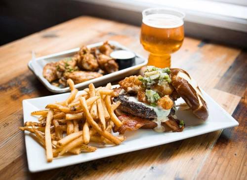 A plate of fries and a sandwich with toppings, alongside a bowl of wings and a glass of beer on a wooden table.