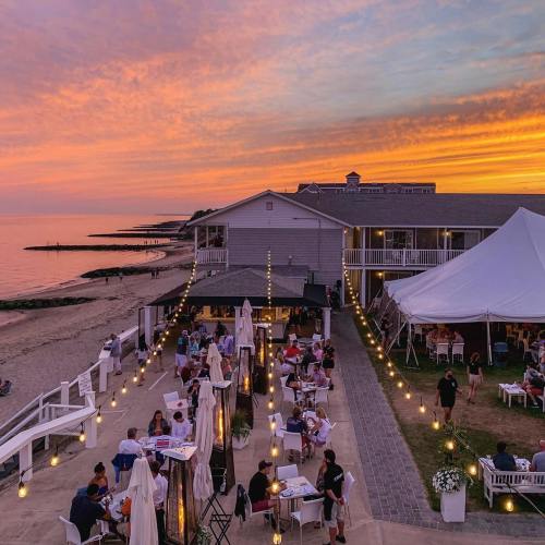 A beachside restaurant at sunset, with diners enjoying outdoor seating under string lights and a colorful sky.