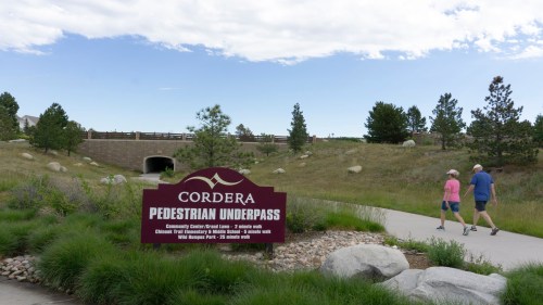 Two people walk along a path near a sign for the Cordera Pedestrian Underpass, surrounded by greenery and blue skies.