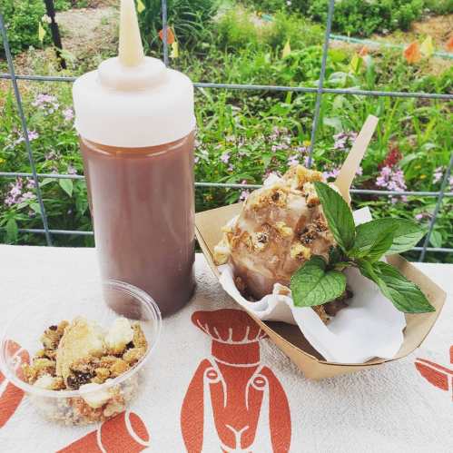 A bottle of sauce next to a dessert topped with nuts and mint, with a small cup of toppings on a patterned tablecloth.