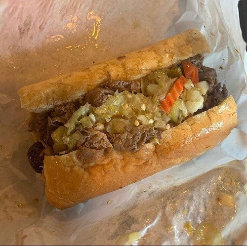 A loaded sandwich with shredded beef, pickles, tomatoes, and onions in a soft hoagie roll, resting on parchment paper.