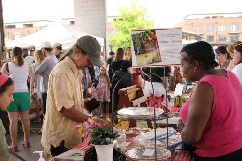 A busy farmers market scene with vendors, shoppers, and colorful displays of fresh produce and goods.