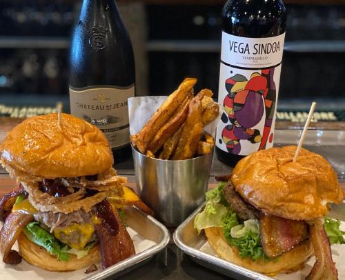 Two gourmet burgers with bacon and toppings, served with a side of sweet potato fries and bottles of wine in the background.