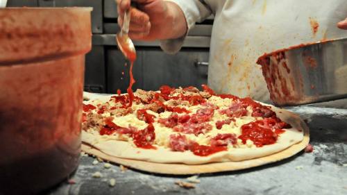 A chef adds sauce and toppings to a pizza dough on a countertop, with ingredients in bowls nearby.
