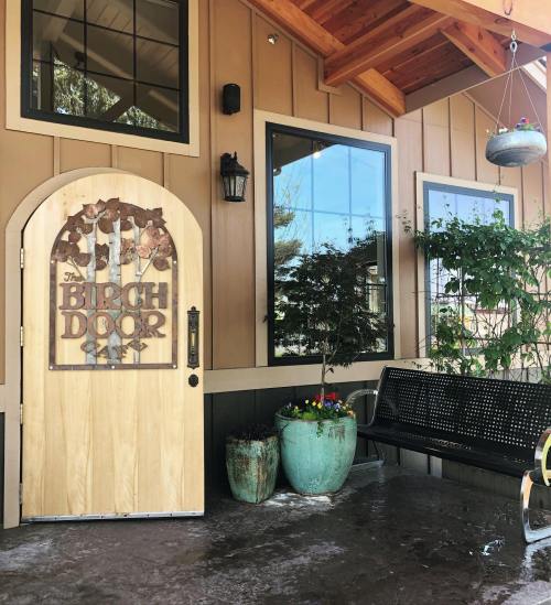 A wooden door with a sign reading "The Birch Door" beside large windows and potted plants on a porch.